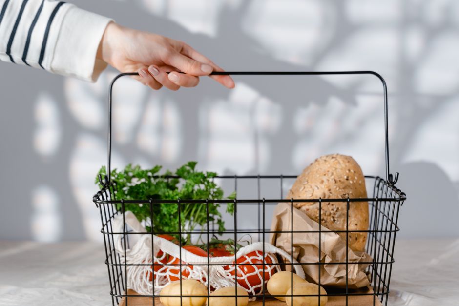 A hand holding a metal basket filled with eco-friendly groceries like bread, vegetables, and parsley.